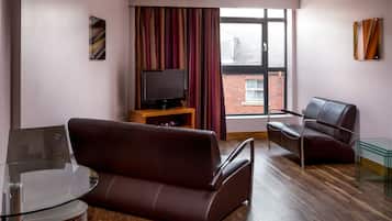 Een moderne woonkamer met een leren bank, een glazen salontafel, een televisie op een houten dressoir, een fauteuil van leer, en een groot raam met gordijnen.