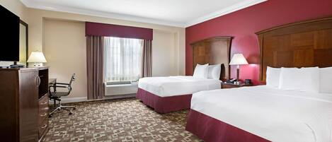 A hotel room with two beds, a desk, a chair, a dresser, and a large window with curtains.