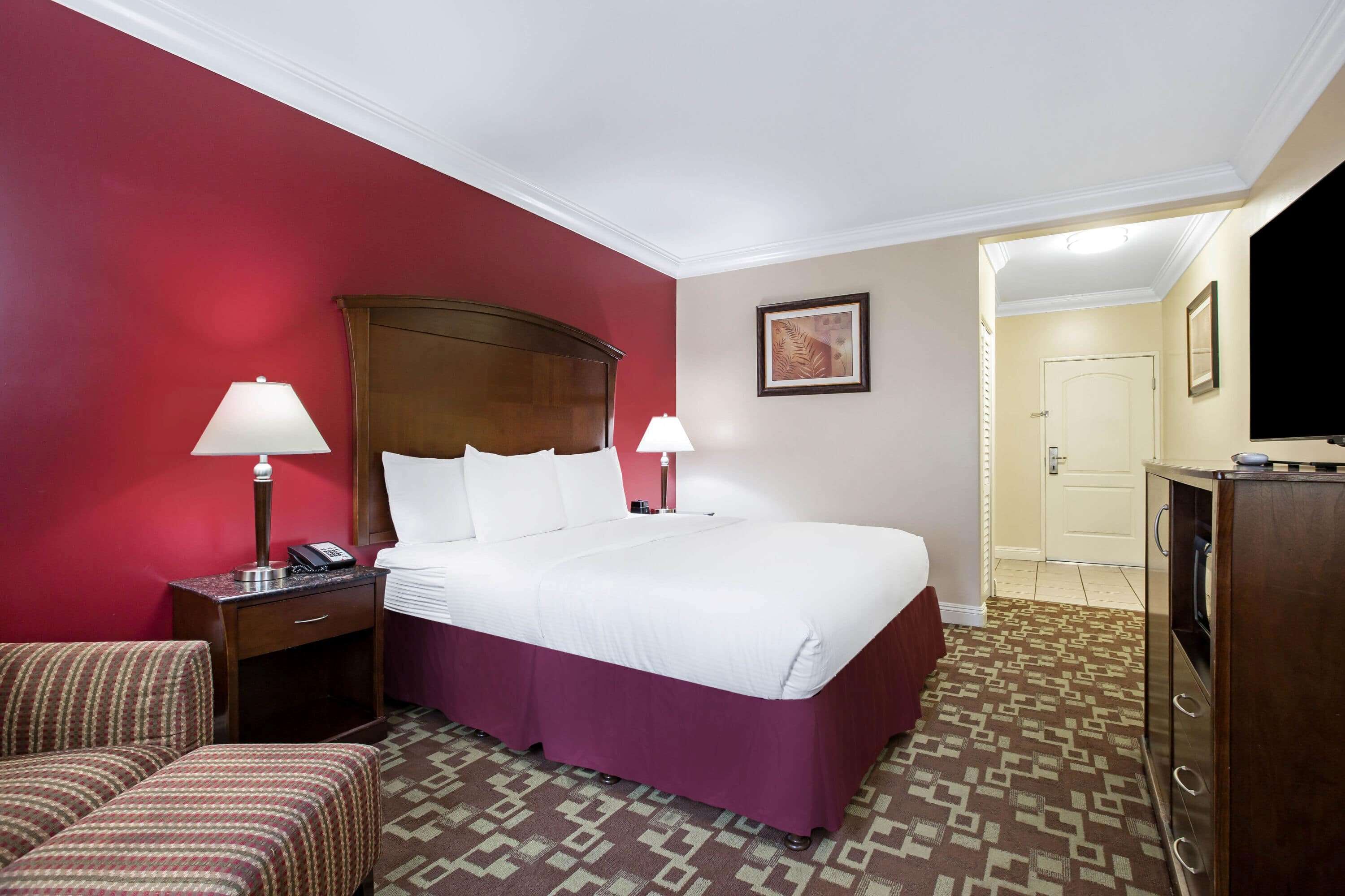 A hotel room with a large bed, two bedside lamps, a wooden headboard, a patterned carpet, a small desk with a lamp, a television, and a door leading to another room.