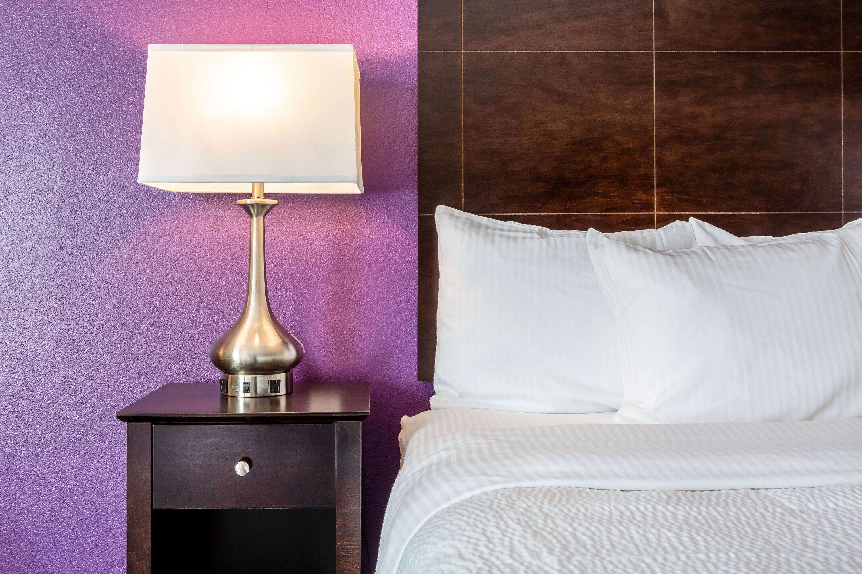 A neatly made bed with white linens, a bedside lamp on a dark wooden nightstand, and a purple wall.