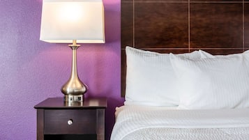 A neatly made bed with white linens, a bedside lamp on a dark wooden nightstand, and a purple wall.