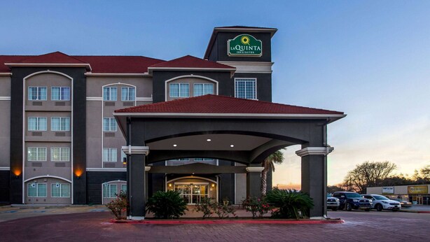 A La Quinta Inn & Suites building with a covered entrance, multiple windows, and a parking lot with cars. - La Quinta Inn & Suites by Wyndham Columbus TX (Columbus)
