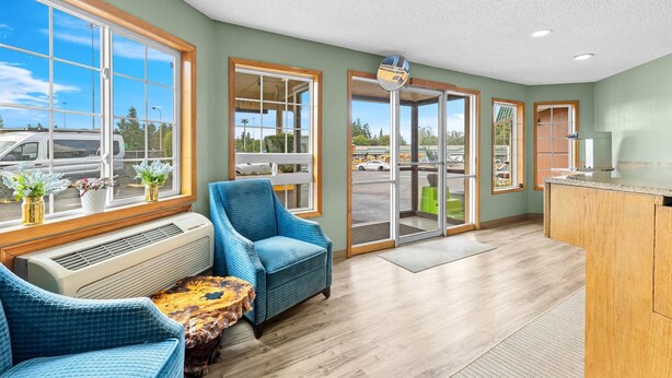 A spacious room with large windows, a blue armchair, a wooden table, and a view of the parking lot. - Rodeway Inn & Suites Portland West - Beaverton (Portland)