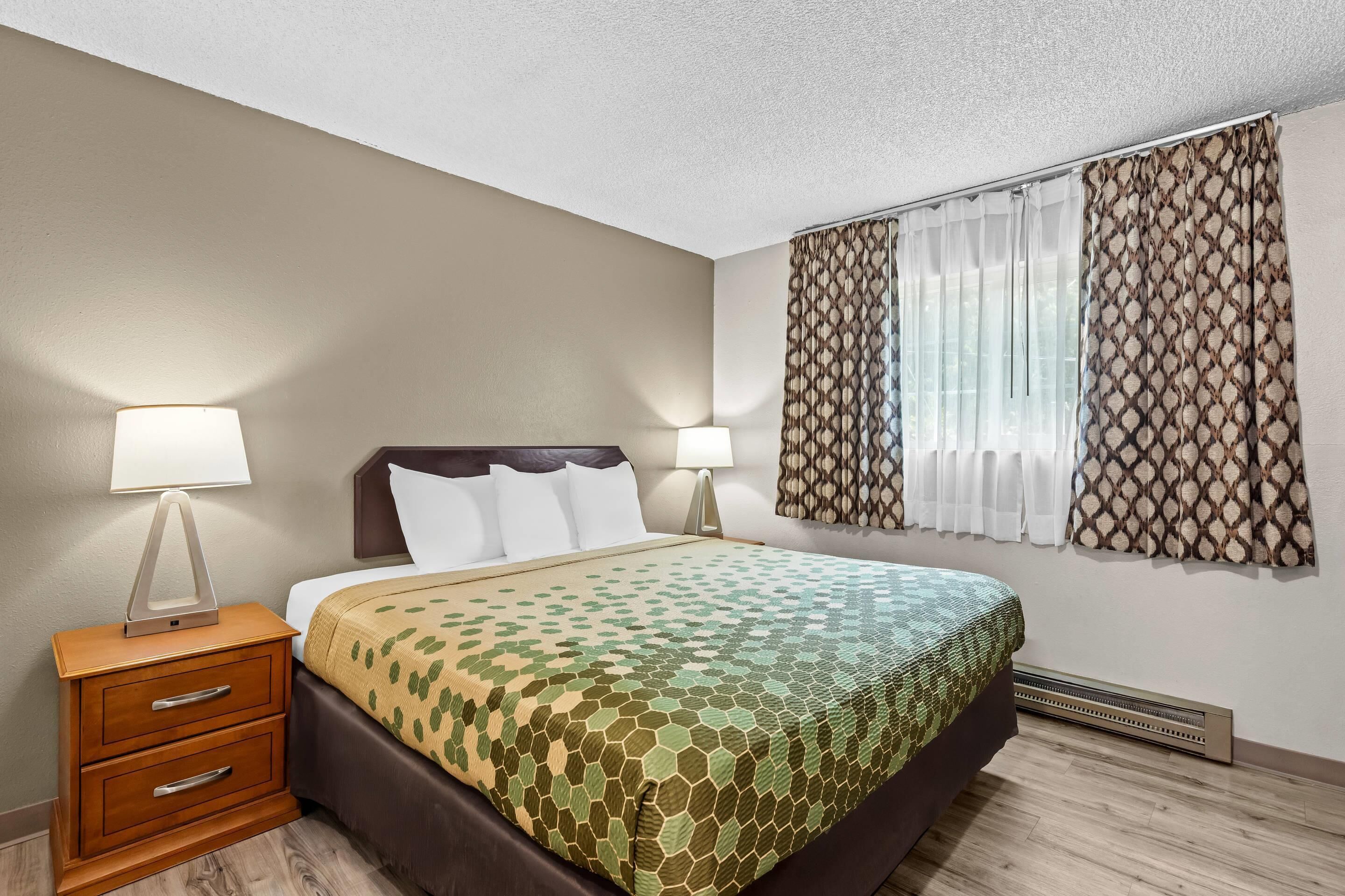A hotel room with a bed, nightstands, lamps, a window with curtains, and a wall-mounted heater.