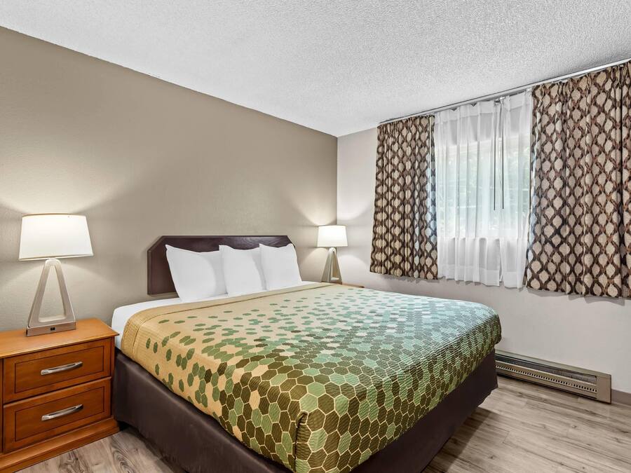 A hotel room with a bed, nightstands, lamps, a window with curtains, and a wall-mounted heater.