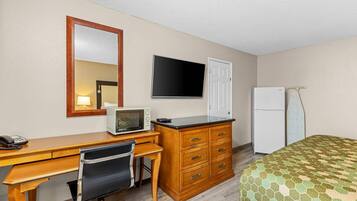 Quarto de hotel com escrivaninha de madeira, cadeira, televisão, frigorífico, cama com colcha estampada e porta.