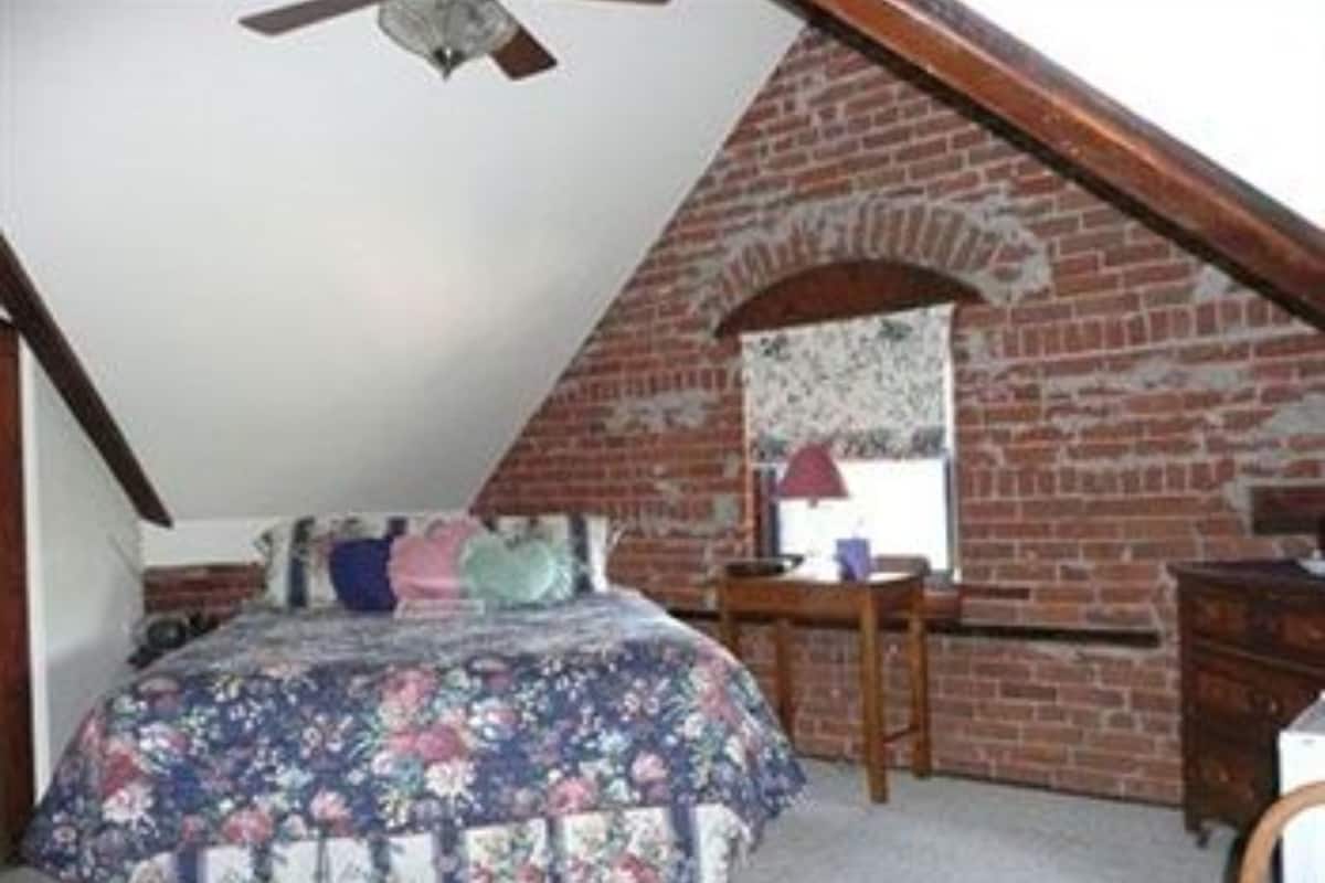 Une chambre avec un plafond en pente, un lit recouvert d’une couvre-lit à motifs floraux, une commode et un miroir accroché au mur de briques.