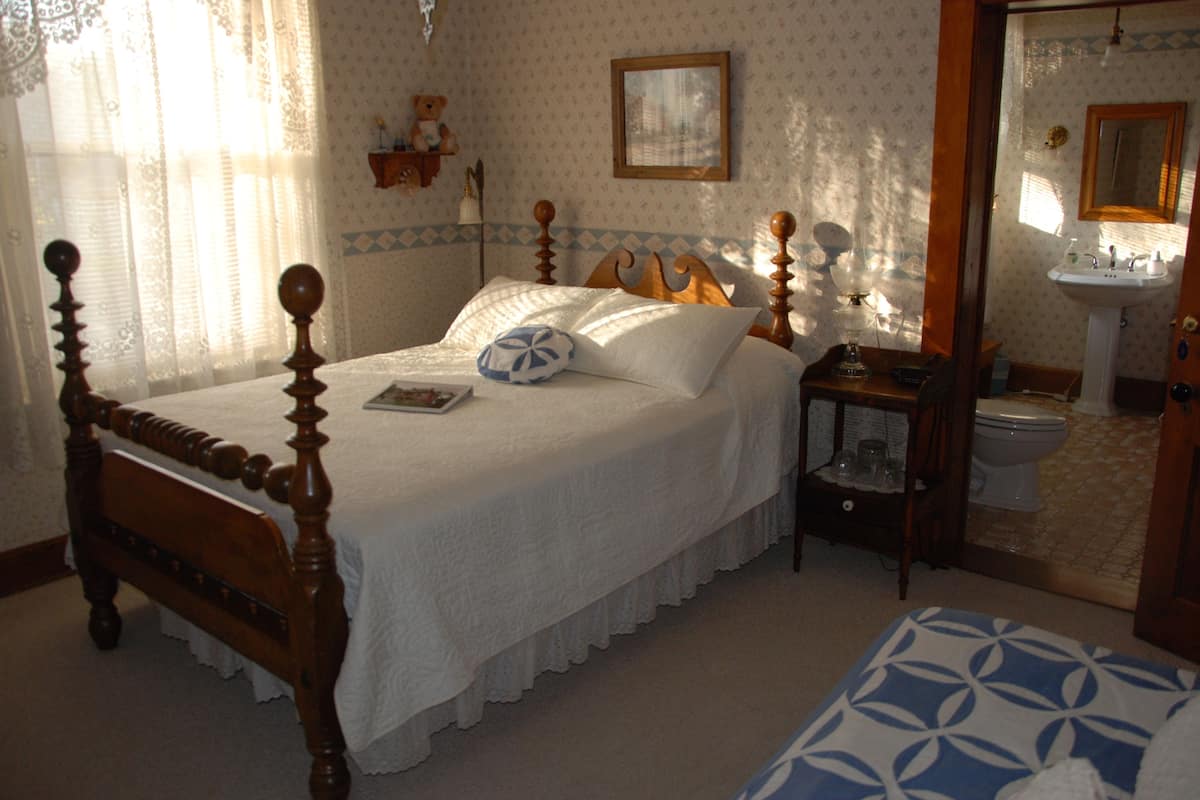 Une chambre à coucher avec un lit à baldaquin en bois, une table de chevet avec une lampe, une petite étagère avec un ours en peluche, et une salle de bain visible à travers une porte ouverte.
