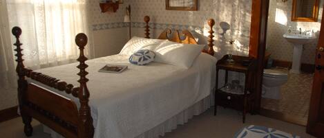 A bedroom with a wooden four-poster bed, a bedside table with a lamp, a small shelf with a teddy bear, and a bathroom visible through an open door.