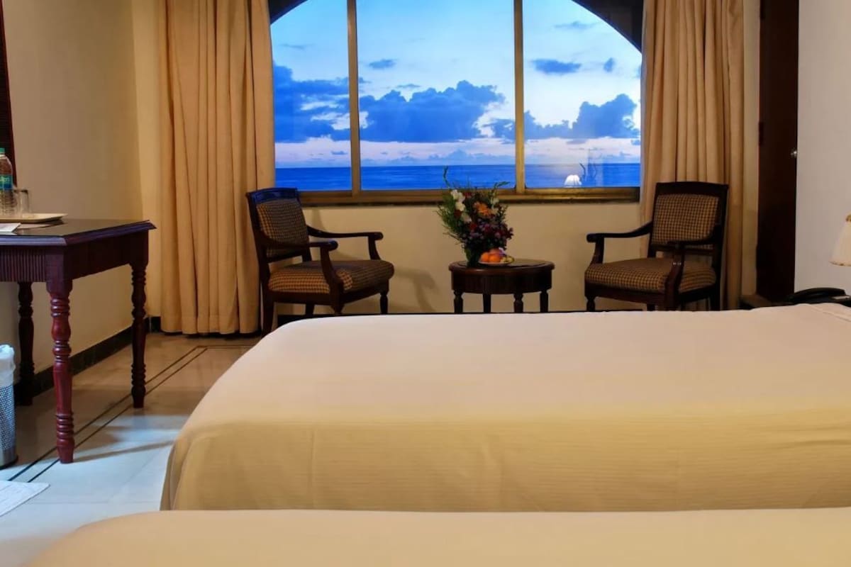 A hotel room with two beds, a desk, and chairs facing a large window with a view of the sea.