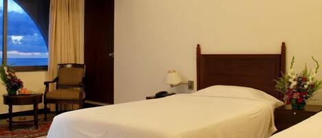 A hotel room with two beds, a chair, a nightstand with a lamp, and a window with a view of the ocean.