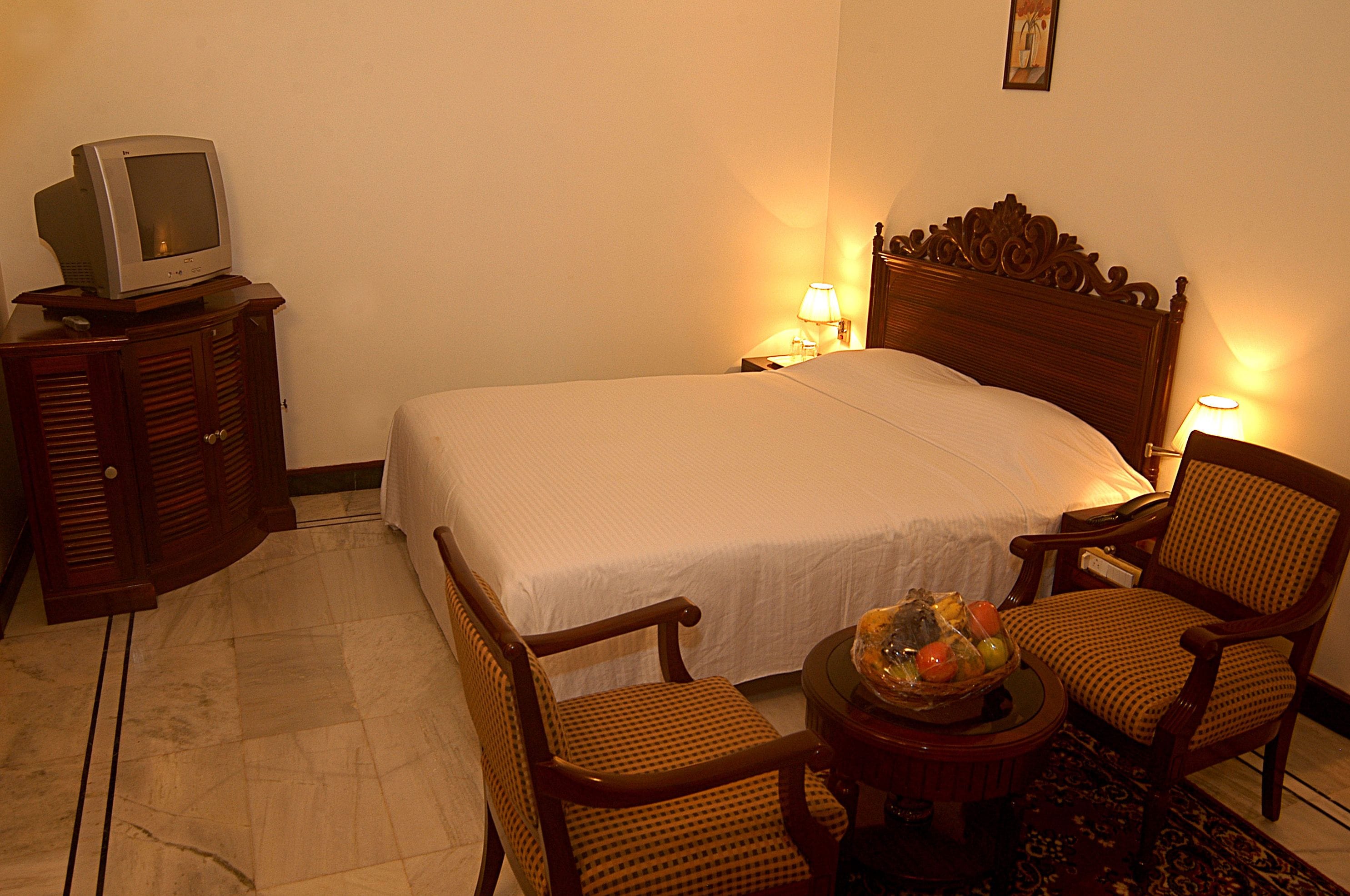 A hotel room with a bed, two chairs, a small table with a fruit basket, a television on a cabinet, and two bedside lamps.