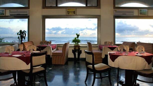A dining area with tables set for two, facing a large window overlooking the ocean. - Paradise Isle Beach Resort (Udupi)