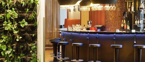 Zona de bar con mostrador curvo, varios taburetes, lámparas colgantes y una pared con plantas trepadoras.
