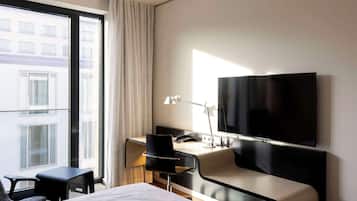 A modern hotel room with a bed, a desk with a chair, a flat-screen TV, and a large window offering a city view.
