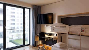 A hotel room with a large window, a desk with a chair, a bed with a ladder, and a TV mounted on the wall.