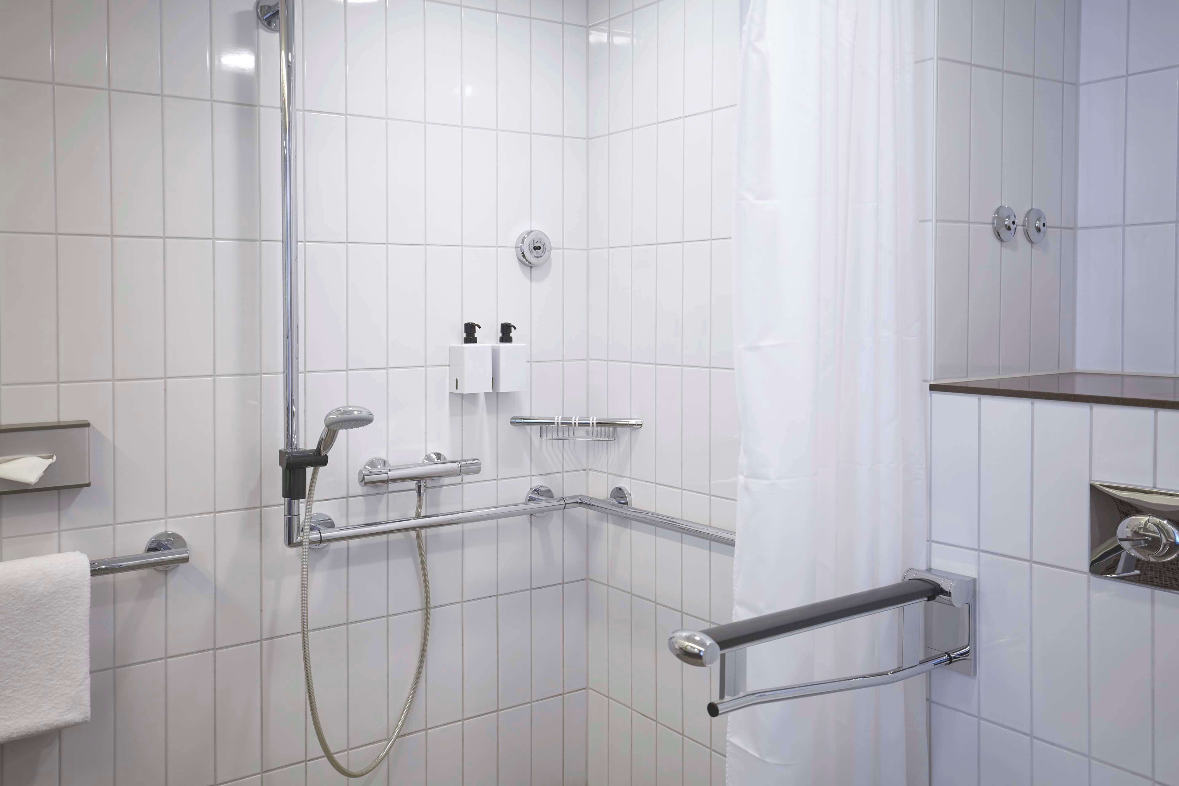 A modern bathroom with white tiles, a shower with a handheld showerhead, a grab bar, and a towel rack.