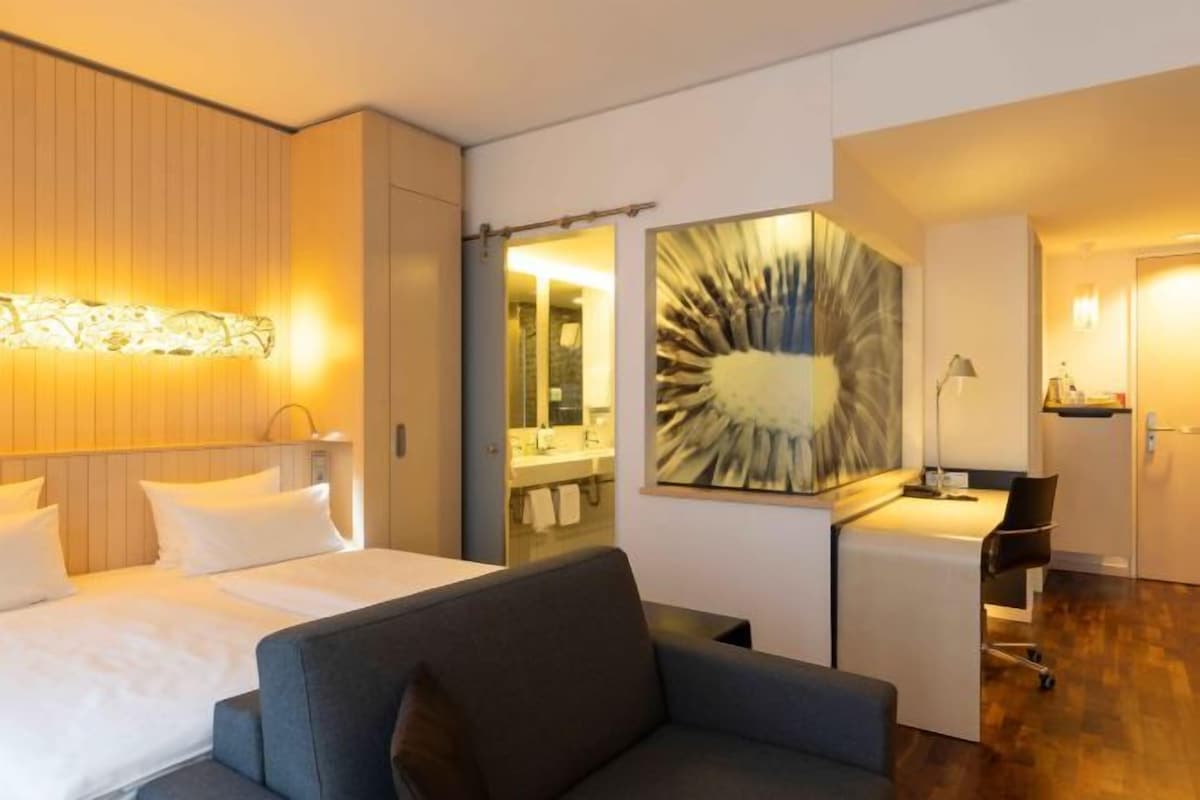 A hotel room with a bed, a sofa, a desk, and a bathroom visible through a glass door.