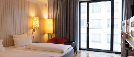 A hotel room with a large bed, a desk, a chair, a lamp, and a view of the city through a large window.