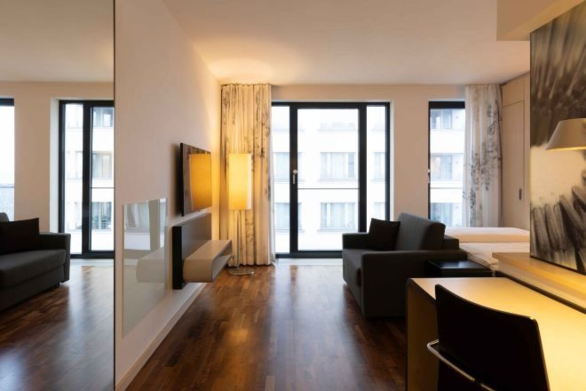 A modern hotel room with a large window, a sofa, a desk, and a lamp.