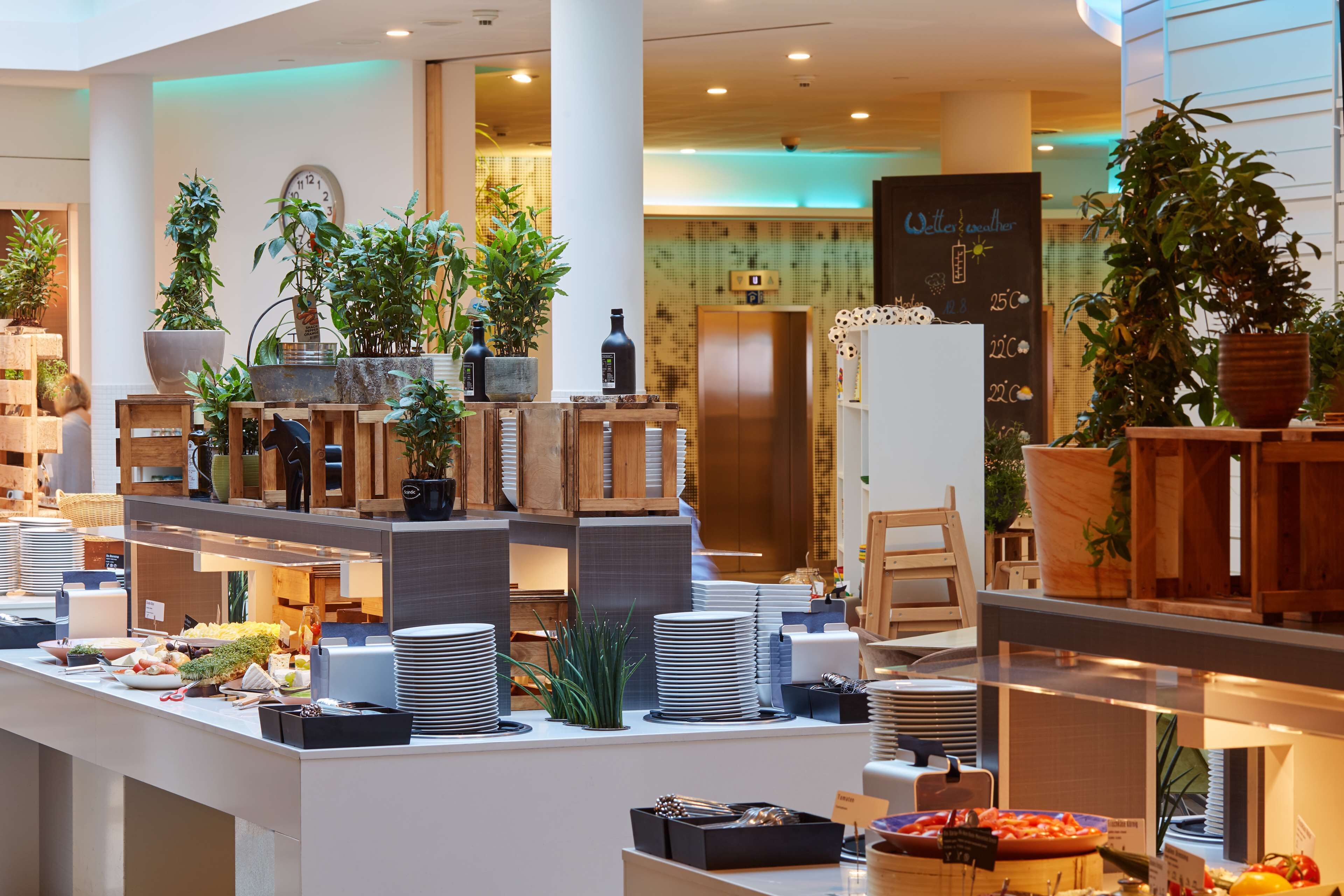 Un hall d’hôtel avec un comptoir d’accueil, de nombreuses plantes en pot, une horloge et un tableau noir affichant les tarifs des chambres.