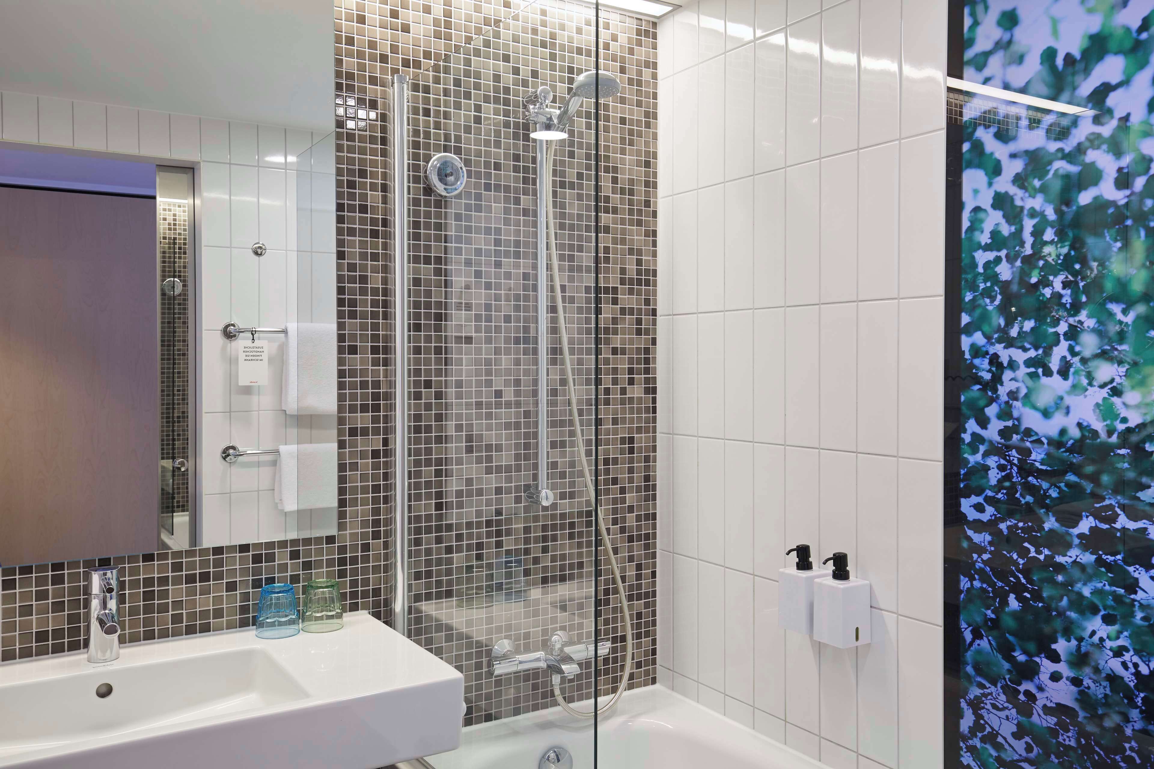 A bathroom with a glass-enclosed shower, a sink with a mirror, and a tiled wall with a nature-themed mural.
