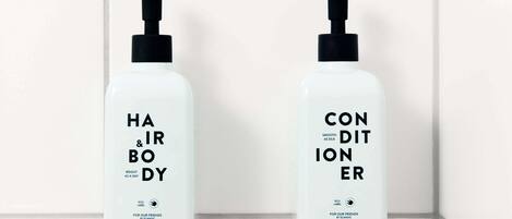 Two white bottles with black pumps, labeled "HAIR & BODY" and "CONDITIONER," hanging on a white tiled wall.