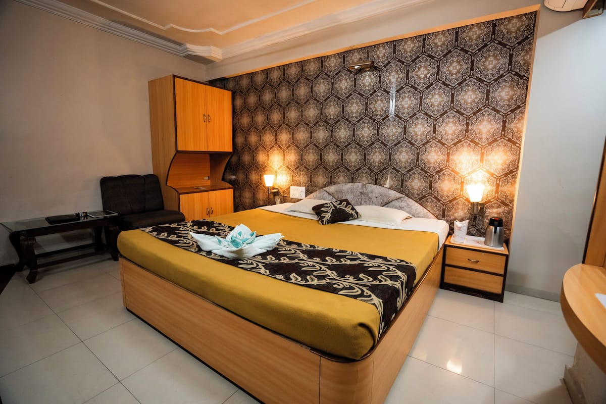 A bedroom with a large bed, a wooden wardrobe, a bedside table with a lamp, and a nightstand with a coffee maker.