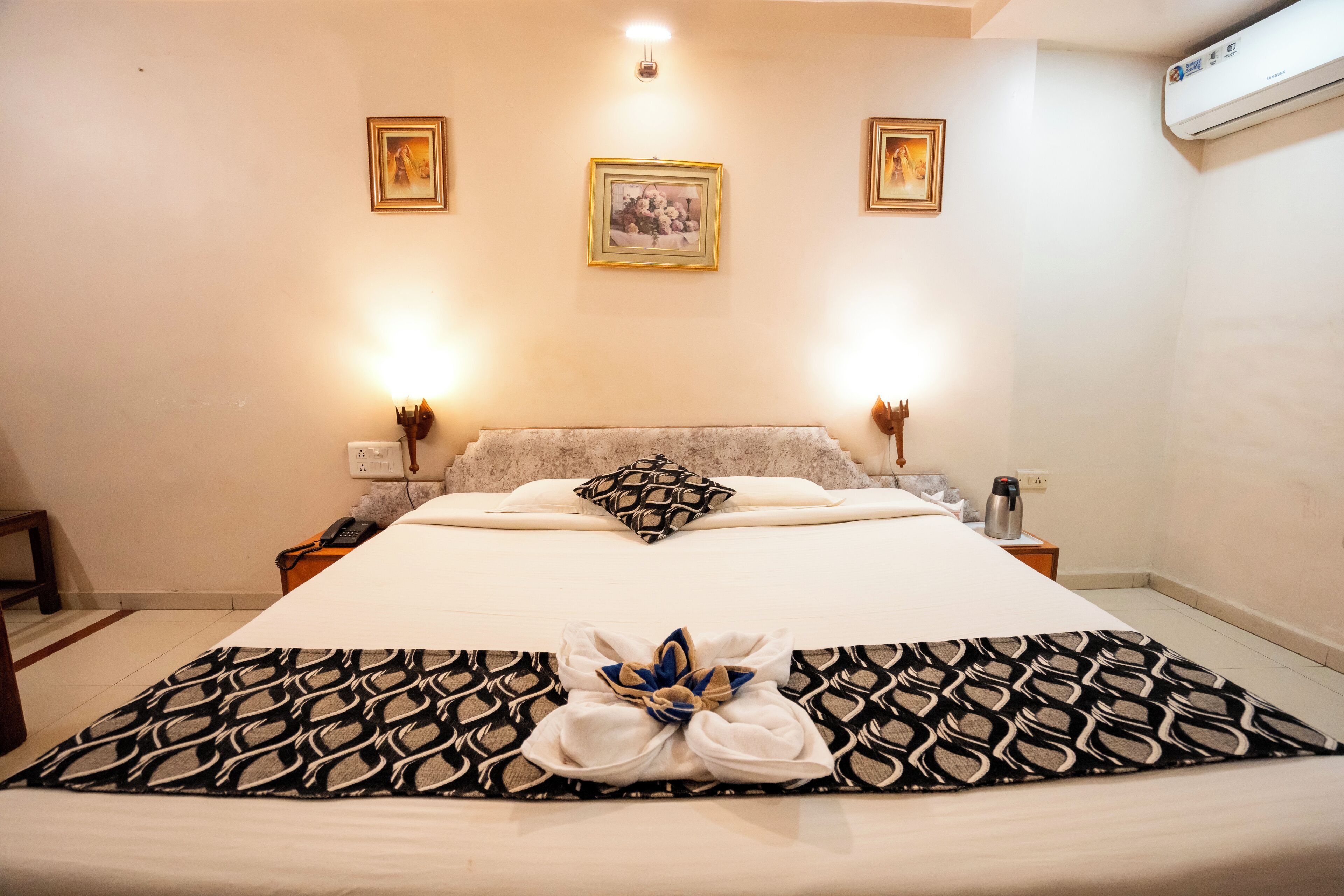A hotel room with a large bed, two bedside lamps, a wall-mounted air conditioner, and framed pictures on the wall.