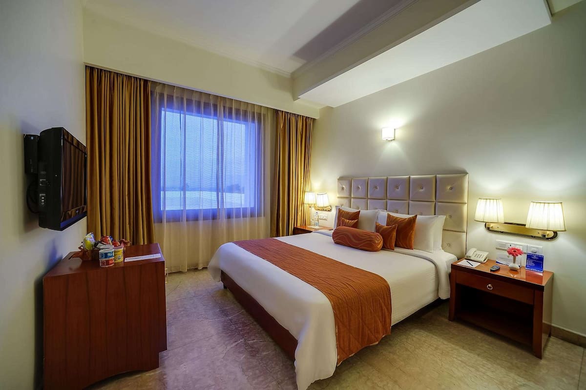 A hotel room with a large bed, a desk with a telephone, a TV, and a window with curtains.