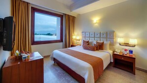 A hotel room with a large bed, a TV, a desk with a lamp, and a window with curtains. - Royal Sarovar Portico Siliguri (Jalpaiguri)