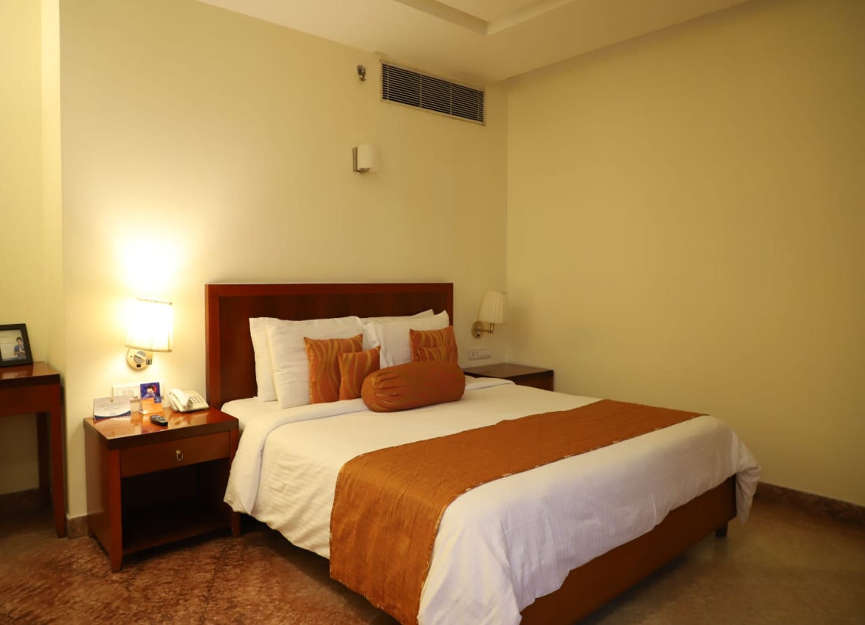 A hotel room with a large bed, a bedside table with a lamp, a phone, and a small table with a picture frame and a book.