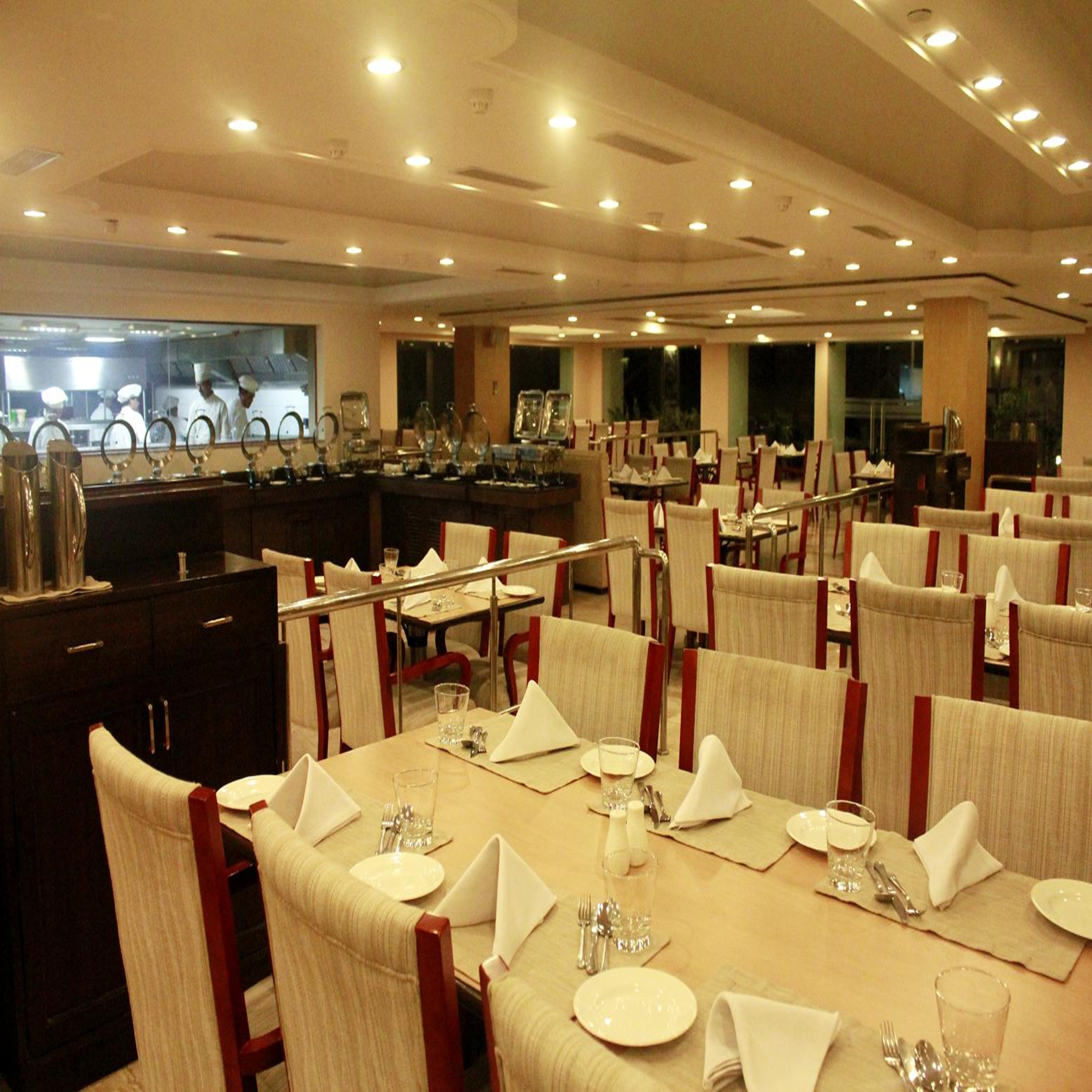 A restaurant with multiple tables set for dining, a kitchen area visible in the background, and a ceiling with recessed lighting.