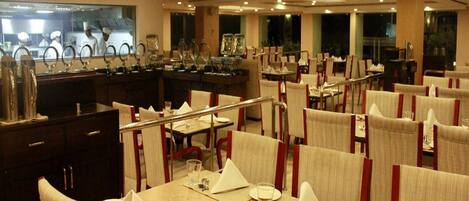 A restaurant with multiple tables set for dining, a kitchen area visible in the background, and a ceiling with recessed lighting.