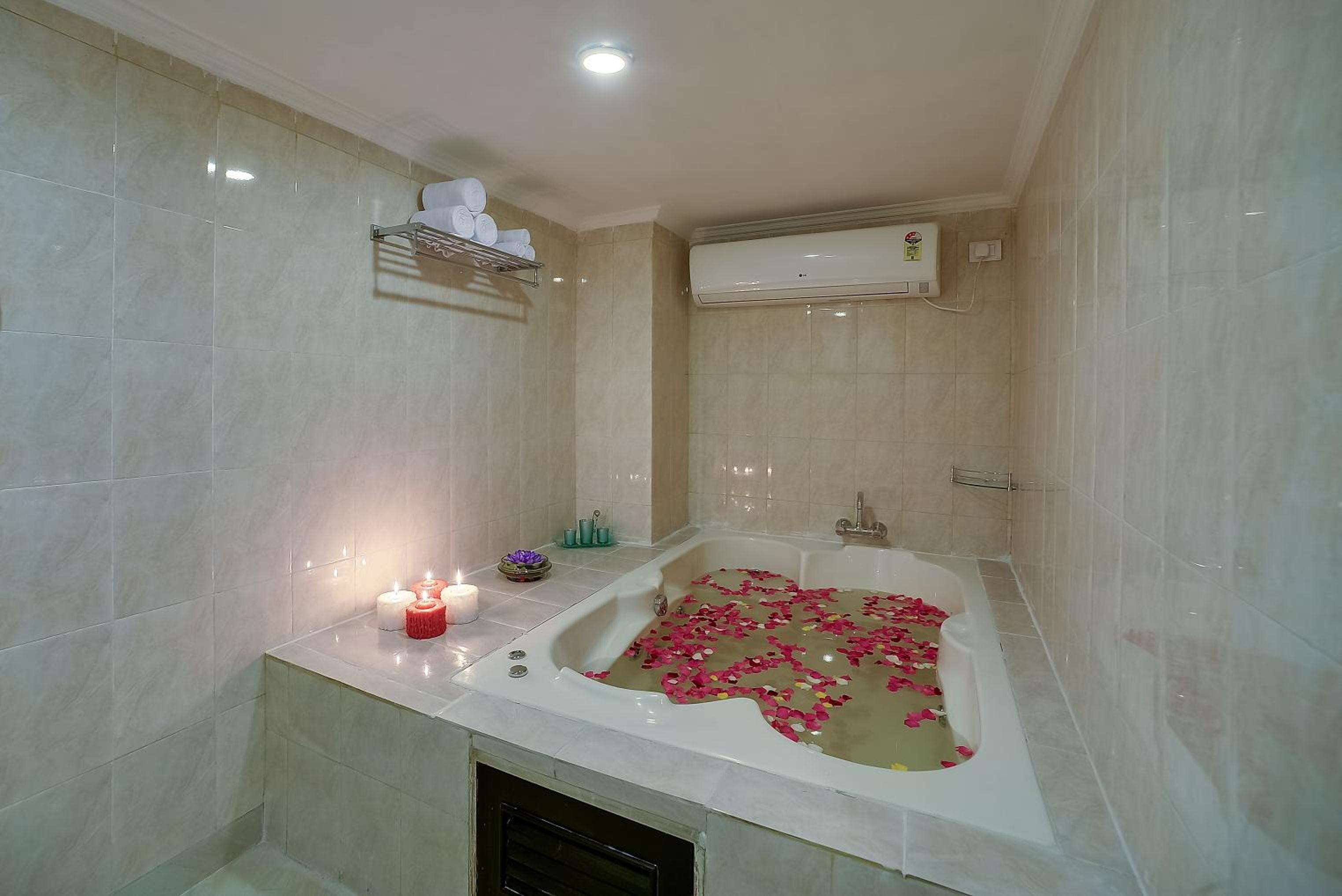 A bathroom with a bathtub filled with rose petals, a lit candle, and a shelf with towels.