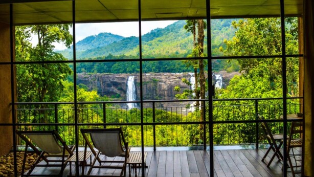 A room with a large window overlooking a waterfall and lush greenery. There are two wooden chairs and a small table. - Rainforest Boutique Resort (Chalakudy)