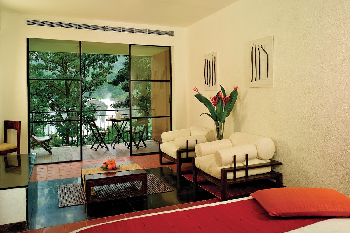A modern room with a large sliding glass door leading to a balcony, two white armchairs, a small table with a vase of flowers, and a red cushion on the floor.