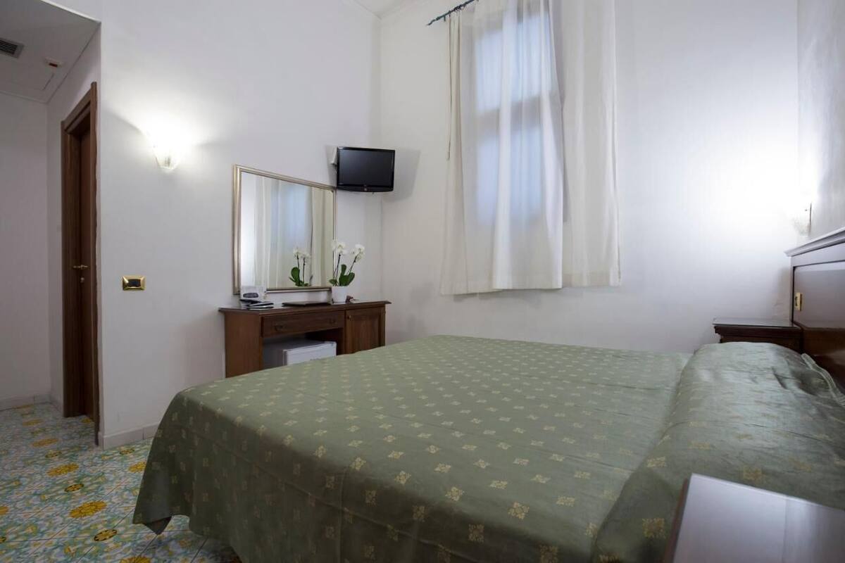 Une chambre à coucher avec un lit, une commode, un miroir, une télévision et une fenêtre avec des rideaux.