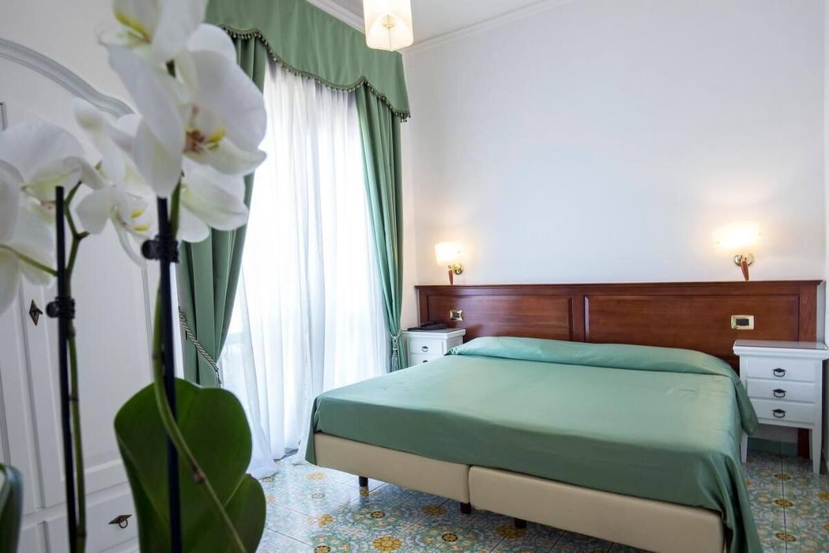 Une chambre d’hôtel avec un lit, des tables de chevet, une tête de lit en bois, un rideau vert et une grande orchidée en pot.