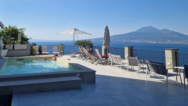 A rooftop pool area with lounge chairs and umbrellas, overlooking a coastal city and a mountain.