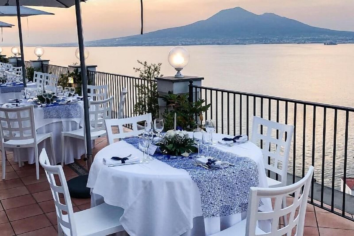 Un espace de restauration en plein air avec des tables et des chaises blanches, une lampe décorative, et une vue sur une étendue d’eau et une montagne en arrière-plan.