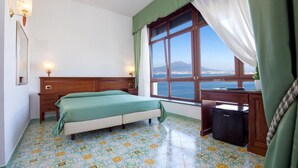 A room with a bed, a desk, a small trash bin, and a window with a view of the sea and a mountain.