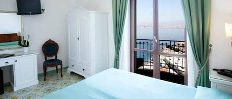 A hotel room with a bed, a desk, a chair, a TV, and a balcony with a view of the sea.
