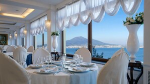 A dining area with tables set for a meal, large windows with a view of the sea and a mountain, and white decor.