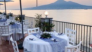 An outdoor dining area with tables set for two, white chairs, and umbrellas. A mountain and body of water are visible in the background.
