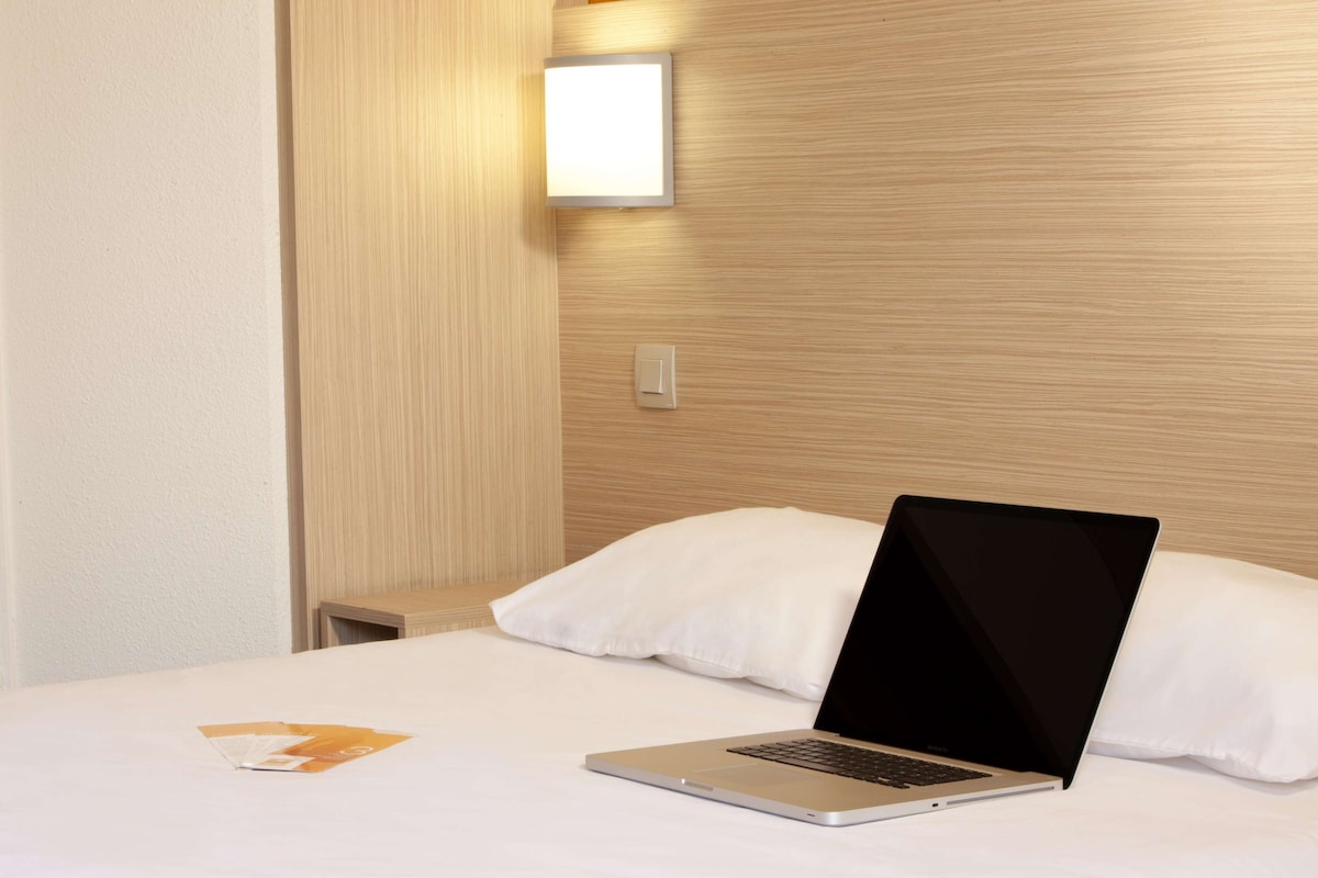 Quarto de hotel com cama, um laptop sobre a cama, mesa de cabeceira e luz acoplada à parede.