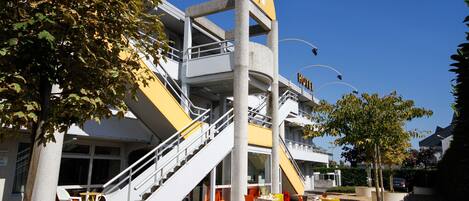 A hotel with a modern design, featuring a central staircase, outdoor seating area with yellow and orange chairs, and a clear blue sky.