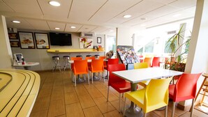 A brightly lit indoor dining area with orange and yellow chairs, a white table, and a counter with a menu display.