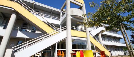 A multi-story building with a yellow and white color scheme, featuring a central staircase and outdoor seating area with yellow and orange chairs.