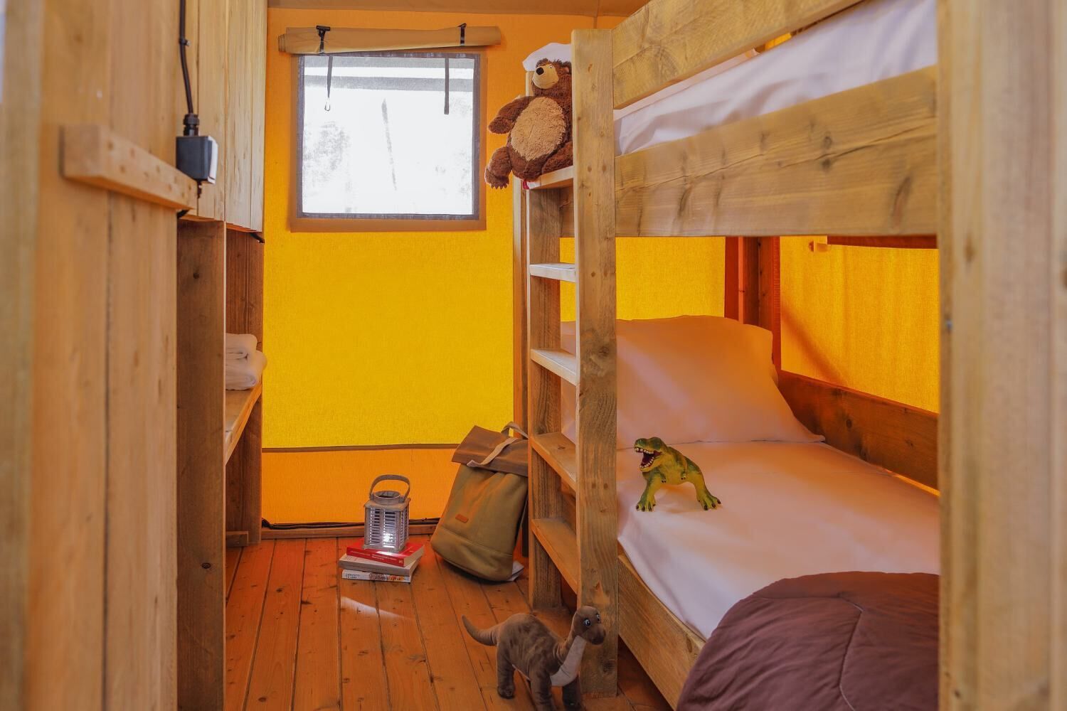 A small, cozy room with a bunk bed, a window with a view, a stuffed bear, a toy dinosaur, and a lantern on the floor.